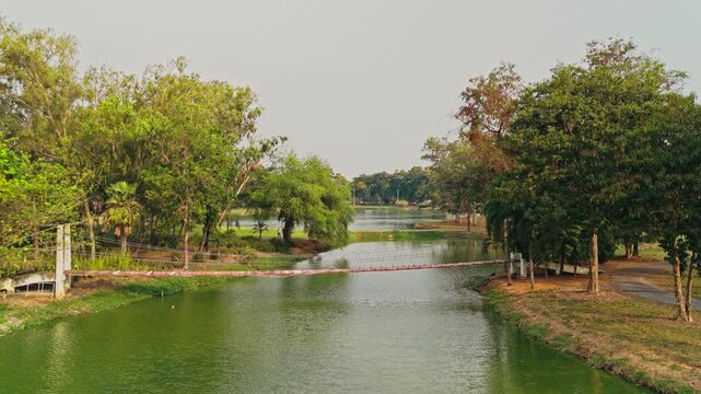 Wooden suspension bridge with red planks hangs suspended above murky green lake water bordered thick leafy trees casting shadows onto grassy shorelines beneath hazy overcast skies approaching aerial