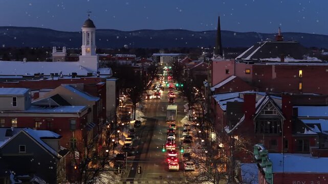 Snowy winter night in downtown Carlisle Pennsylvania with cars moving along main street glowing lights church tower and mountains in background showing small town life of United States. aerial view.