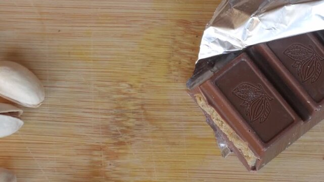Chocolate bar partially unwrapped on wooden board near pistachios