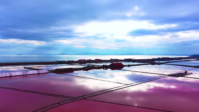 Landscape view of salt evaporation pans with mirror reflections of clouds and coastal buildings on the horizon. Vibrant pink salt ponds reflecting cloudy sky at sunset.