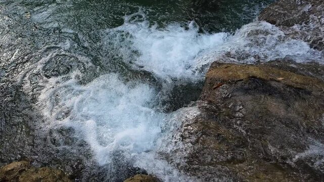 Cinematic slow motion of crystal clear water flowing over rocks in a natural stream. Bubbling white foam and emerald ripples splashing in a tropical forest waterfall. High quality 4K nature background
