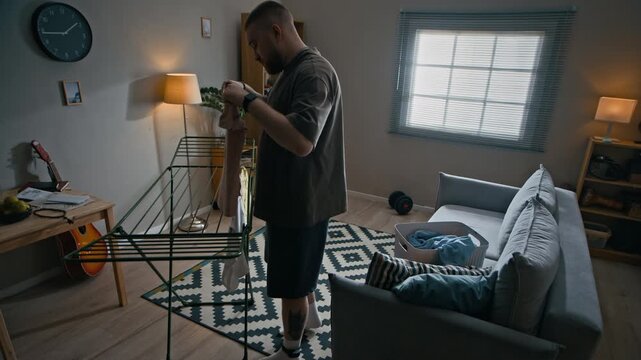 Full shot of Caucasian man under house arrest hanging clothes on indoor drying rack in apartment while completing daily household chores