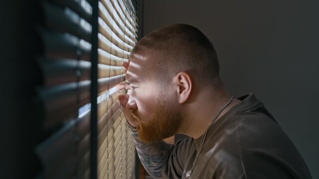 Side view medium closeup of perplexed Caucasian bearded man approaching window and peeking through blinds with caution and concern