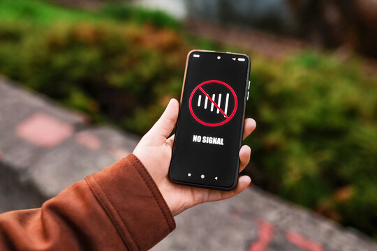 Hand holding a smartphone displaying a "No Signal" icon with a crossed-out network bar outdoors. Concept of poor cellular connection, internet outage, or lack of coverage.