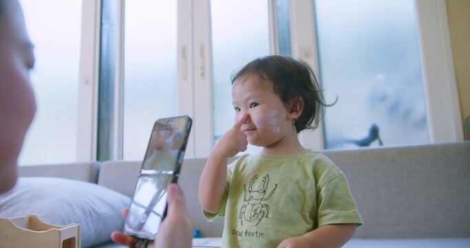 An adorable toddler with face paint playfully poses for a smartphone camera as a parent records a video for social media content creation in a modern home.