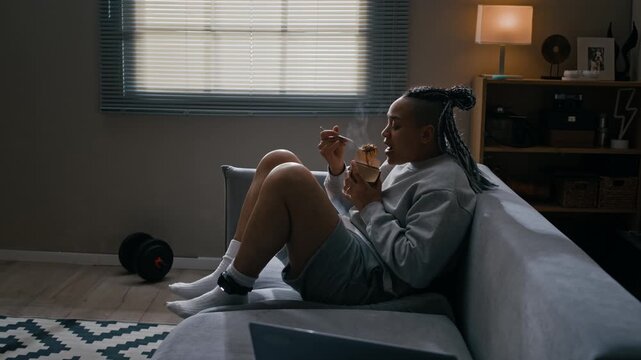 Side view shot of Biracial woman with electronic monitoring bracelet sitting on sofa and eating takeout noodles from box in apartment