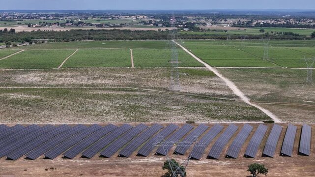 Aerial view of a solar farm with rows of photovoltaic panels and high voltage power lines across green agricultural fields in Palmela, Setubal, Portugal.