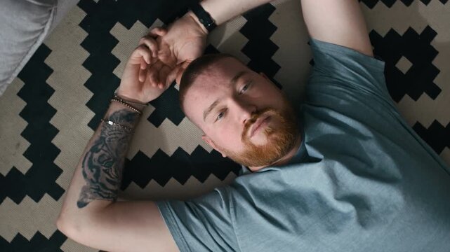 Arc shot of tattooed and bearded man lying on patterned rug with hands behind head at home, looking frustrated while reflecting on personal problems and life decisions