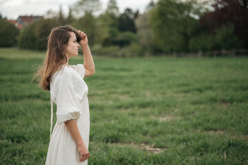 Obraz premium Romantic young woman touching hair in green field, dreamy outdoor portrait with soft summer light.