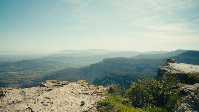 A rocky ledge rises above a sweeping mountain landscape, with steep drop-offs, layered ridges, and distant valleys fading into soft atmospheric haze in Catalonia, Spain.