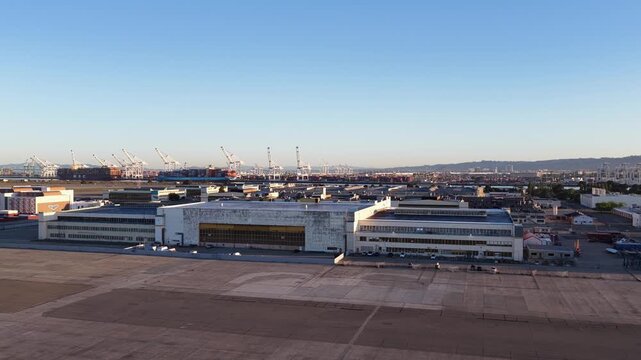 Expansive aerial view slowly panning over the former naval air station in Alameda, California, revealing the industrial landscape and the busy port of Oakland container terminal in the background