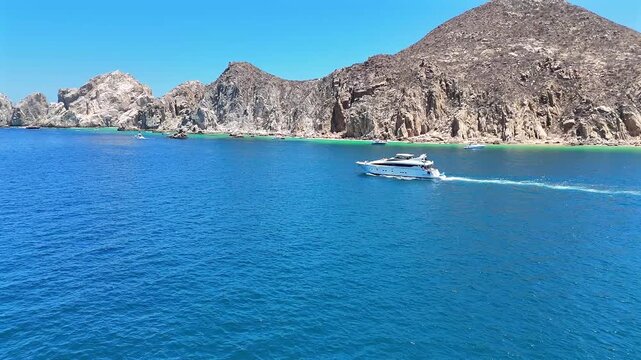 Drone closing in on a yacht heading toward El Arco de Cabo San Lucas, with the final stretch of coastline and beaches behind it.