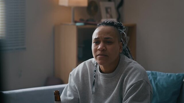 Tilt down shot of Biracial woman wearing electronic ankle monitor drinking beer and watching television while relaxing on couch in apartment during home confinement