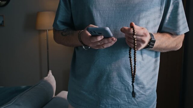 Tilt up cropped shot of unrecognizable man wearing electronic ankle monitor holding smartphone and prayer beads at home during house arrest, reflecting stress and personal coping