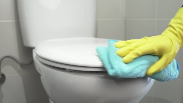 Close up of hand in a yellow rubber glove with blue rag cleaning the toilet in the bathroom. Disinfect and sanitary standards.Scrubbing and cleaning.