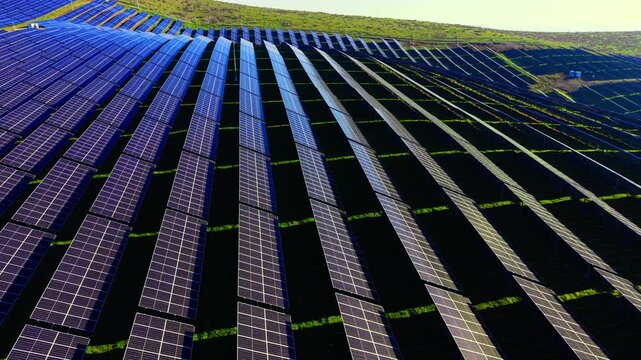 Aerial perspective of repetitive photovoltaic array lines following the terrain of green grass hills. Geometric patterns of solar panel rows on rolling hills.