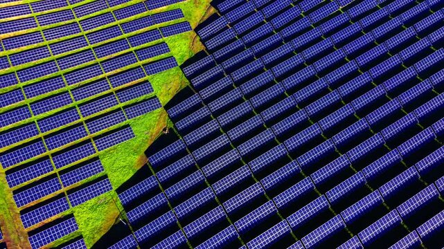 Contrast between blue photovoltaic panels and green grass patches. Aerial view of solar panel arrays on terrain.