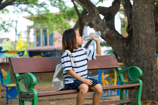 Cute Asian student girl sitting on a park bench and drinking water from a plastic bottle, Back to school and healthy lifestyle concept.