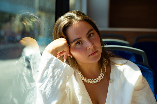 A young woman wearing a white shirt and pearl necklace sits inside a bus leaning her head on her hand near the window.