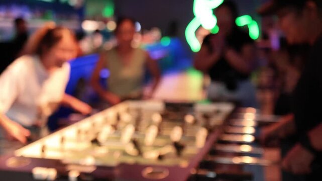 Blurred footage of young adults playing table football game and celebrating goal at indoor entertainment center with neon lights.