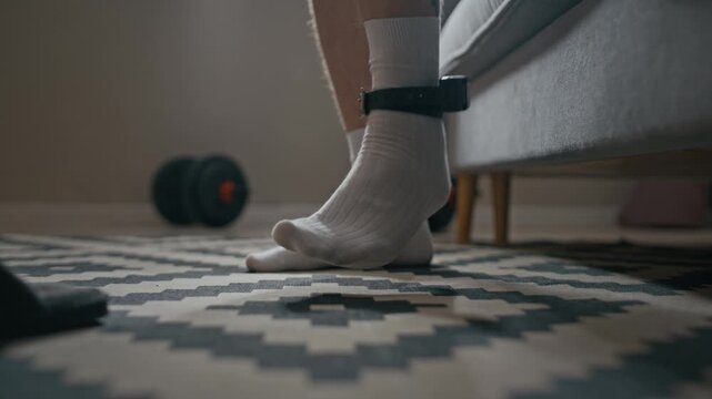 Low section shot of unrecognizable person with tattooed ankle wearing monitoring device and slippers while getting up from sofa