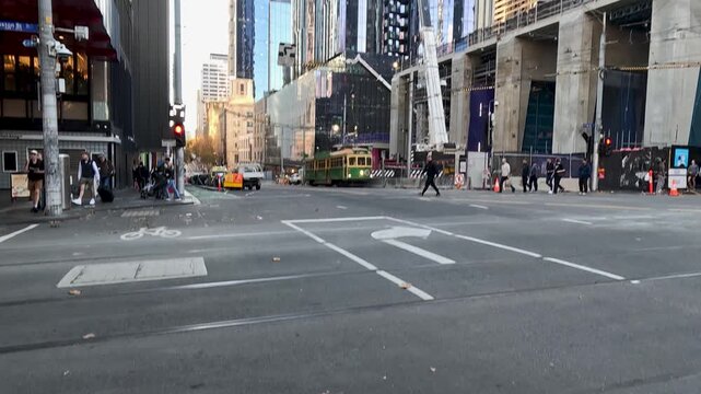 Pedestrians and vehicles at busy intersection in Melbourne city center with modern blue skyscraper and vintage green tram.