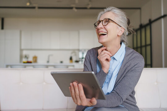 Happy senior woman laughing while using tablet on sofa at home, elderly lifestyle with technology, online communication, digital connection, leisure time and modern retirement concept