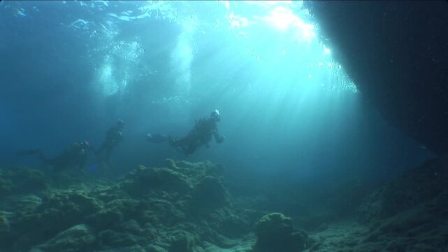  scuba divers exploring around a reef underwater deep blue water big rocks and bubbles ocean scenery 