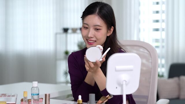 Asian beauty influencer applying makeup using compact mirror filming product review tutorial for social media content creation at home living room