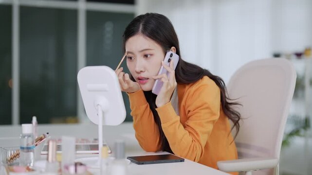 young Asian women applying makeup cosmetics using smartphone mirror beauty routine preparing morning skincare tutorial tutorial at home modern interior bright
