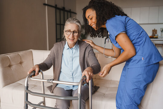 Young female caregiver assisting elderly woman with walker to stand up at home, senior patient receiving mobility support, healthcare help and rehabilitation concept