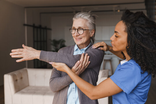 Young female nurse helping senior woman stretch her arm at home, caregiver supporting elderly patient during rehabilitation exercise, mobility recovery, home care service and healthcare concept