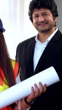 Indian Asian group of civil engineers and architects wearing fluorescent safety jackets, hard hats, holding rolled blueprints, standing confidently with folded arms, smiling, looking at camera