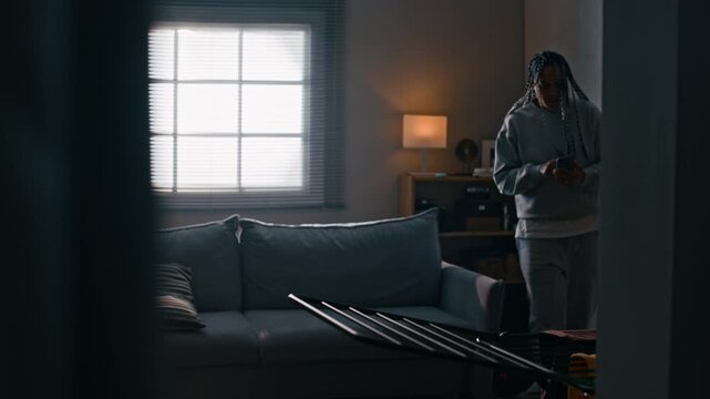 Wide shot of Biracial woman with braided hair wearing electronic ankle bracelet pacing around apartment while checking smartphone messages then sitting down on sofa
