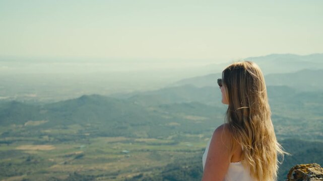 A close view of a young woman in sunglasses gazing across a hazy mountain valley, with soft light, distant ridges, and a quiet sense of elevation in Catalonia, Spain.