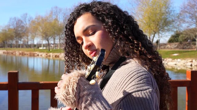 Young woman with curly hair playing the ukulele with a lake in the background on a sunny day. Close up
