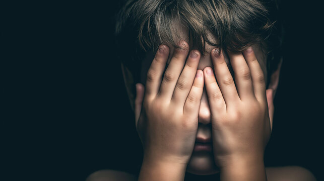Child hiding face with hands, featuring dramatic low key lighting and a dark background, depicting emotions of fright and stress