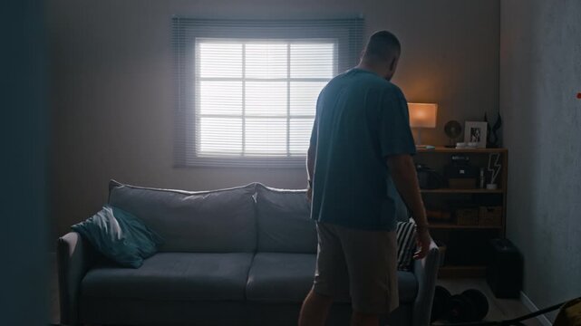 Track side view shot of depressed man wearing electronic ankle monitor walking back and forth in apartment, using smartphone and throwing darts while feeling bored and frustrated during house arrest