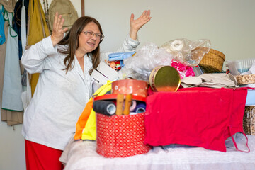 Stressed Middle aged woman with glasses raises hands in frustration standing before pile cluttered items, Home organization sorting household items, Cleaning routine clutter lifestyle transformation © kittyfly