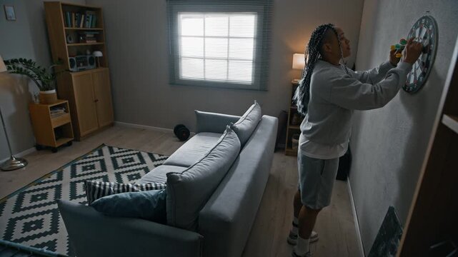 Wide shot of Biracial woman wearing electronic ankle monitor throwing darts in apartment while passing time during house arrest and home confinement