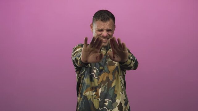 Man soldier in camouflage shirt shields eyes with one hand and reaches forward with the other in a studio pink backdrop, grimacing in discomfort; defense distress.