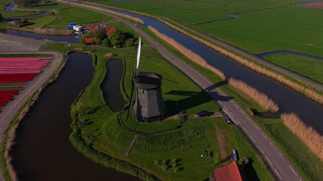 Aerial view of the historic Bovenmolen G windmill standing by a canal amidst lush green polder fields under bright sunlight Schermerhorn, Noord-Holland, Netherlands.