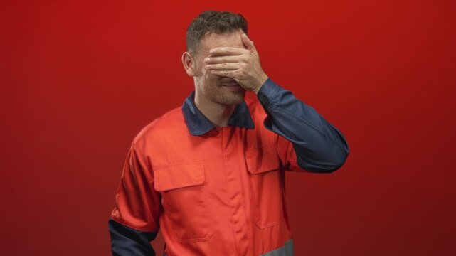Young hispanic man in orange coverall with blue sleeves covers eyes with hand in studio against red backdrop; frustration fatigue isolation.