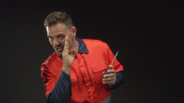 Man handyman in orange uniform cups hand to mouth, holding screwdriver and smiling in studio; helpful repair service.