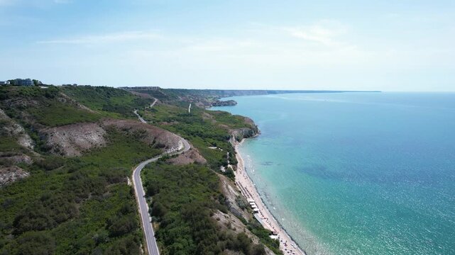 Aerial view of a winding coastal road along green cliffs and a sandy beach by the turquoise sea in Balchik, Dobrich Province, Bulgaria.