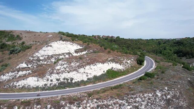 Aerial view of a winding asphalt road curving around white limestone cliffs and green hillsides with a cyclist in Balchik, Dobrich Province, Bulgaria.