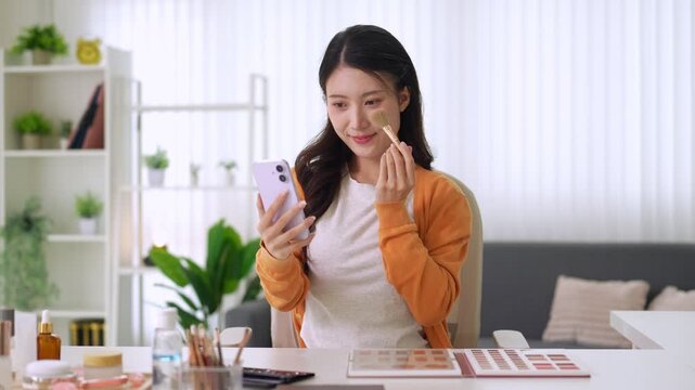 young Asian women beauty influencer recording makeup tutorial video using smartphone applying foundation brush cosmetic product live streaming online at home