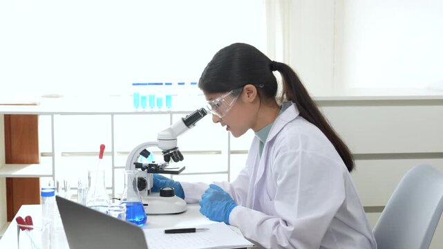 Young Asian women scientist working with test tube in laboratory. Scientist in a lab coat and safety glasses handles a test tube at a desk in a laboratory, make note on laptop, experimentation in a co