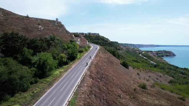 Aerial view of cyclists riding along a winding coastal road overlooking the Black Sea and rocky cliffs in Balchik, Dobrich Province, Bulgaria.