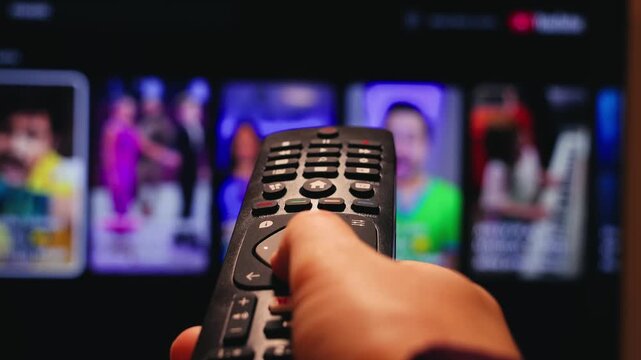 A close-up of a hand holding a TV remote control and pointing it at the screen. A blurred TV interface with streaming content appears in the background. Conceptually, this refers to home entertainment